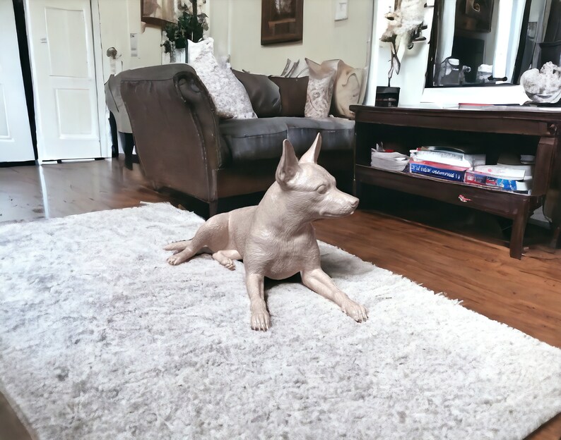 Puede incluir: Una figurita de perro de cer&aacute;mica de color marr&oacute;n claro tumbada sobre una alfombra blanca y esponjosa. El perro est&aacute; en una pose relajada con la cabeza girada hacia un lado.