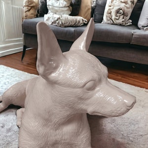 Puede incluir: Una escultura de cer&aacute;mica rosa claro de un perro acostado, con la cabeza girada hacia un lado. El perro tiene un acabado liso y brillante y un pelaje detallado.