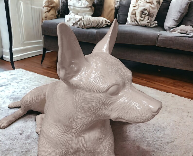 Puede incluir: Una escultura de cer&aacute;mica rosa claro de un perro acostado, con la cabeza girada hacia un lado. El perro tiene un acabado liso y brillante y un pelaje detallado.