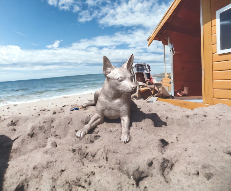 Puede incluir: Una estatua de perro de color plateado tumbada en una playa de arena frente a una casa de playa de madera.
