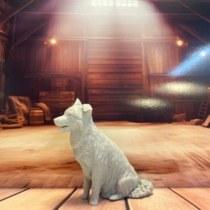 May include: A white ceramic sculpture of a dog sitting on a wooden floor. The dog is looking to the left and has its mouth slightly open. The background is a rustic wooden barn.
