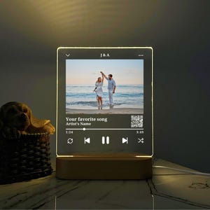 May include: A clear acrylic night light with a wooden base. The light features a photo of a couple on a beach, a music player interface, and the text "Your favorite song Artist's Name".