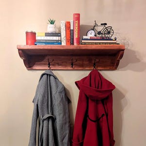 May include: A wooden shelf with a decorative edge, featuring two hooks on the bottom. A gray coat and a red coat are hanging on the hooks. The shelf is decorated with books, a small potted plant, a clock, and a toy bicycle.
