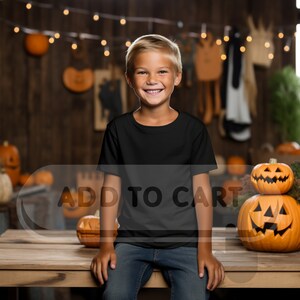 Può includere: Un bambino piccolo con una maglietta nera e jeans blu sorride alla telecamera. È seduto su un tavolo di legno con zucche sullo sfondo. Il testo "ADD TO CART" è visibile nell'immagine.