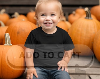 Può includere: Un bambino piccolo con una maglietta nera e jeans blu è seduto su una superficie di legno davanti a un mucchio di zucche arancioni. Il bambino sorride e guarda in alto. Il testo "ADD TO CART" è sovrapposto all'immagine.