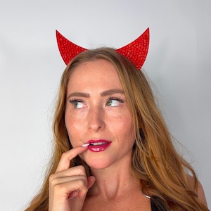May include: Red devil horns with a glittery texture. The horns are attached to a headband. The image is a close-up shot of the horns.