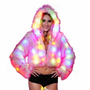 Puede incluir: Una chaqueta de piel sintética rosa con capucha, iluminada por luces LED de colores. La chaqueta tiene un diseño corto y cierre de cremallera. Las luces son en tonos amarillos, verdes y rosas, creando un efecto vibrante.