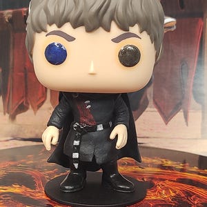 以下が含まれることがあります： 茶色の髪のキャラクターのFunko Popビニールフィギュア。黒い衣装にマントとブーツを着用しています。片方の目は青、もう片方の目は黒です。フィギュアは黒い円形の台座に立っています。