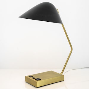 May include: A modern desk lamp with a black, cone-shaped shade and a brass-colored base and arm. The base has three black buttons. The lamp is on a white surface.