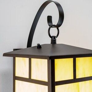 May include: Black outdoor wall sconce with a square, frosted glass shade and a decorative scroll arm. The light is on and the glass is illuminated with a warm yellow glow.