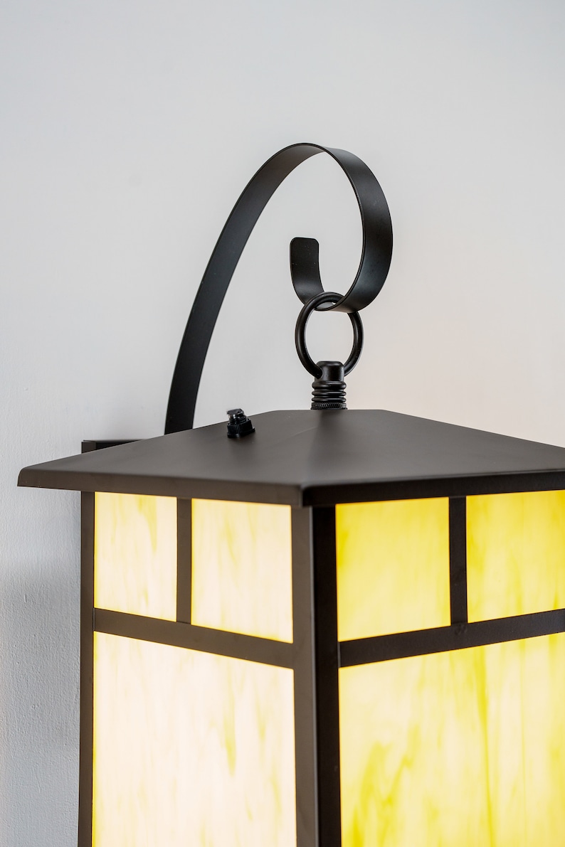 May include: Black outdoor wall sconce with a square, frosted glass shade and a decorative scroll arm. The light is on and the glass is illuminated with a warm yellow glow.