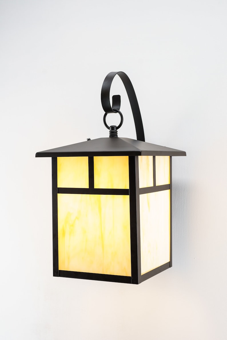 May include: A black metal outdoor wall sconce with a square, frosted glass shade. The sconce has a decorative scroll design on the top.