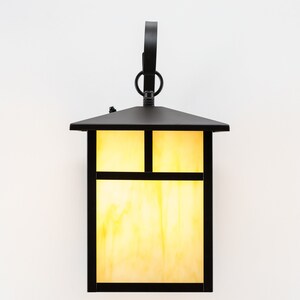 May include: A black metal outdoor wall light fixture with a rectangular glass shade. The glass shade has a grid pattern and is illuminated with a warm yellow light.
