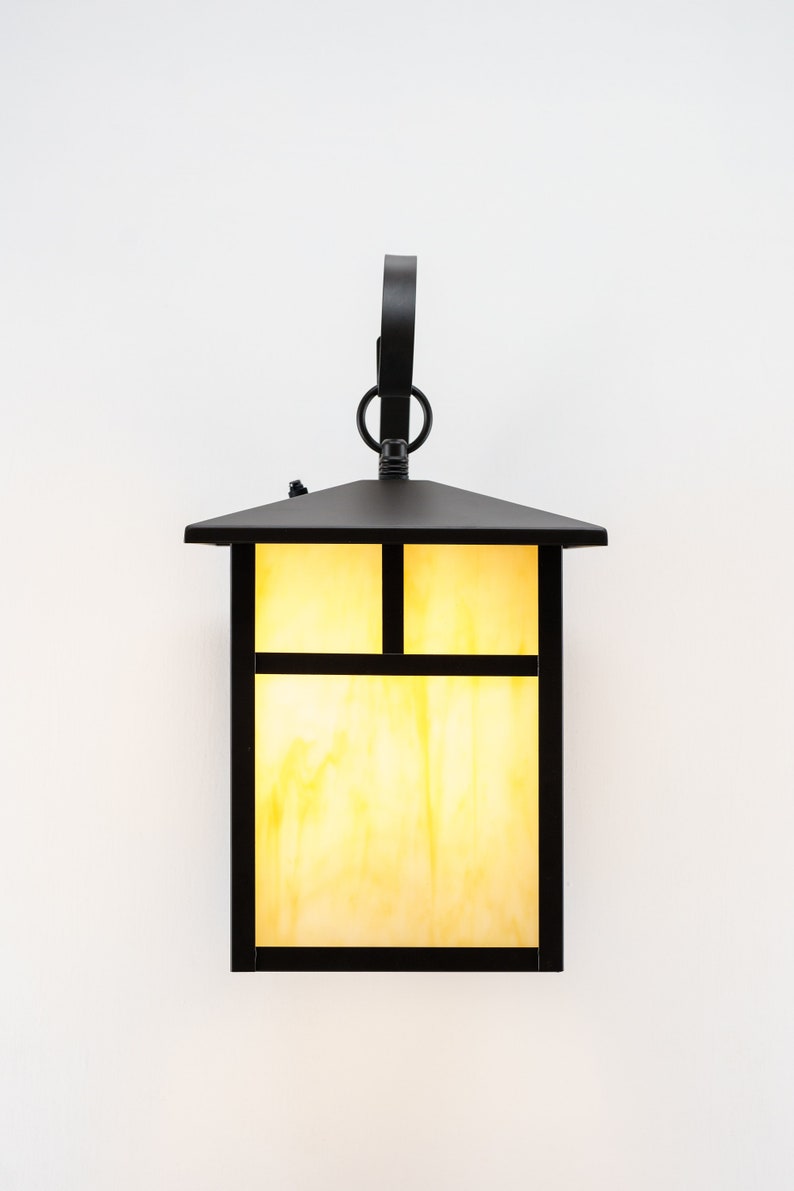 May include: A black metal outdoor wall light fixture with a rectangular glass shade. The glass shade has a grid pattern and is illuminated with a warm yellow light.
