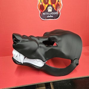 May include: A black, molded mask with white teeth, resembling a canine or wolf. The mask has a strap and eye holes. A sticker with a paw print design and the text "METALHOUND studios" is in the background.