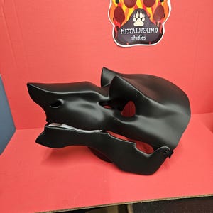 May include: A black, stylized mask with a sharp, angular design. The mask has a prominent snout and open mouth, suggesting a creature or animal. The background is red, with a sticker that says "METALHOUND studios".