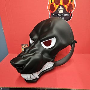 May include: A black wolf mask with white teeth and red eyes. The mask has a menacing expression and is made of a smooth, matte material. The mask is designed with a strap for wearing. A logo with a paw print is visible in the background.