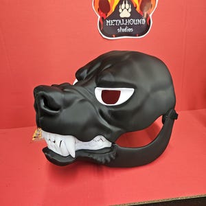 May include: A black dog mask with white teeth and red eyes. The mask has a detailed snout and is designed to fit over the face. The mask is made of a durable material and is likely for cosplay or costume use.