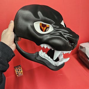 May include: A black panther mask with white fangs and eye sockets. The eyes have a yellow and orange design. The mask is held against a red background.