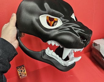 Könnte beinhalten: Eine schwarze Panthermaske mit weißen Zähnen und Augenhöhlen. Die Augen haben ein gelb-oranges Design. Die Maske wird vor einem roten Hintergrund gehalten.