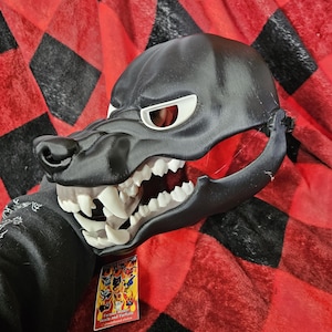 May include: A black and white wolf mask with a toothy grin. The mask is made of plastic and has a realistic look. The mask is on a red and black checkered background.