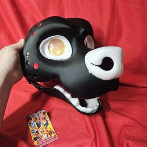 Könnte beinhalten: Schwarze und weiße Fellmaske mit einer weißen Nase und offenem Mund. Die Maske hat ein gelbes Auge mit einem Gittermuster. Die Maske wird von Metalhound Studios hergestellt, einem Hersteller von Fellkostümen für Köpfe und Partials.