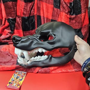 May include: A black and white wolf mask with a detailed mouth and teeth. The mask is made of a smooth, hard plastic and is held in a person's hand. The mask is sitting on a red and black plaid blanket.