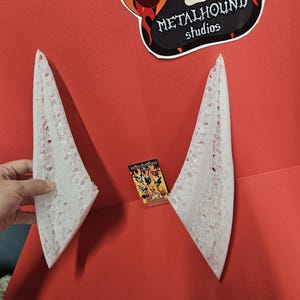 May include: Two white, pointed, decorative objects with a lace-like design are displayed against a red background. A business card with multiple dog illustrations is visible. The Metalhound Studios logo, featuring a paw print, is at the top.