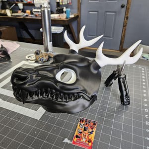 May include: A black dragon mask with white antlers and a toothed jaw. The mask has a large eye opening and is resting on a gray cutting mat. A business card with the text "METALHOUND" is next to the mask.