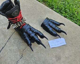 May include: Black leather boot with a matching claw-shaped attachment. The boot features red laces, silver spikes, and a black zipper. A separate claw piece is also visible. The text on the paper reads "June 10, Aaron Moore, Metalhound Studios."