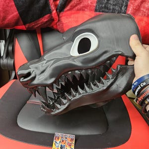 May include: A black and white furry mask with a large open mouth and sharp teeth. The mask is made of a hard material and has a realistic look. The mask is being held by a hand, and there is a red and black plaid blanket in the background. The mask is labeled "Metalhound Studios" and "Fursuit Maker Heads and Partials".