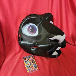 Puede incluir: Una cabeza de fursuit negra con forma de pantera, con ojos grandes y expresivos y dientes blancos. La cabeza tiene varios orificios de ventilación y está hecha de un material liso y brillante. Una pequeña tarjeta de visita con ilustraciones coloridas está al lado.
