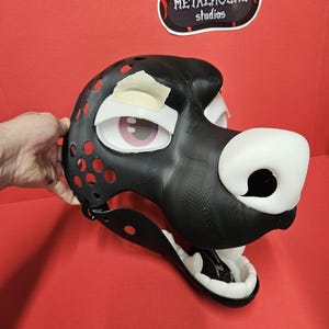May include: A black and white fursuit head with a large white nose and pink eyes. The head has red circular vents and is held by a hand. The logo for Metalhound Studios is visible in the background.