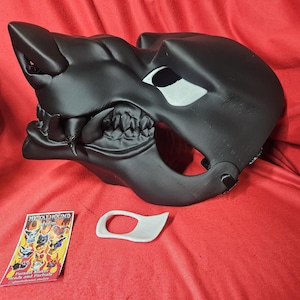 May include: A black wolf mask with a white eye detail. The mask is made of plastic and has a textured surface. The mask is lying on a red surface. The mask is a prop for cosplay or theater.