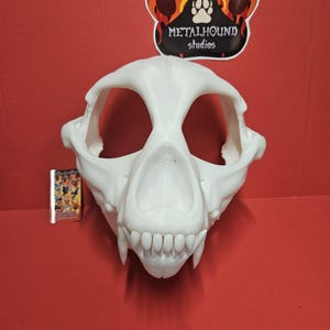 May include: A white, 3D-printed animal skull mask featuring large eye sockets, a prominent snout, and visible teeth. Above the mask is a sticker with the text METALHOUND studios and a paw print icon. A small rectangular card is visible to the left.