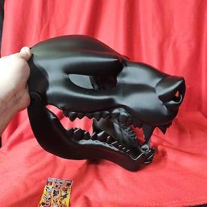 May include: A black 3D printed wolf mask with an open mouth and sharp teeth. The mask is made of plastic and has a realistic design. The mask is on a red surface.