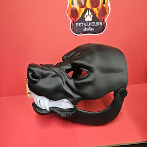 May include: A black dog mask with white teeth. The mask has eye holes and a strap. The Metalhound Studios logo is visible in the background.