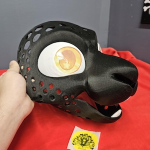 May include: A black, 3D-printed mask with large, round eye sockets featuring yellow and orange mesh. The mask has a snout and is held against a red surface. The mask has several circular holes. A yellow sticker with a graphic is visible.
