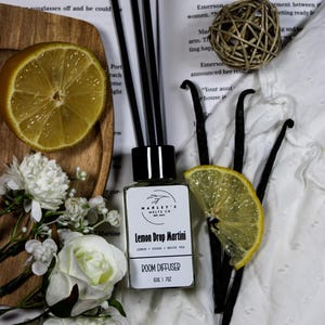 Może przedstawiać: Dyfuzor do pomieszczeń "Lemon Drop Martini" jest prezentowany z plasterkami cytryny, strąkami wanilii i białymi kwiatami. Dyfuzor ma czarne patyczki i czarną nasadkę. Obraz zawiera drewnianą deskę do krojenia i plecioną kulę.