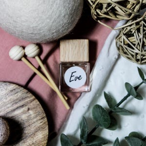 Puede incluir: Naturaleza muerta con un pequeño frasco de perfume con tapa de madera y el nombre "Eve" en una etiqueta blanca. La escena incluye una gran bola blanca, esferas decorativas de madera y ramitas verdes, todo sobre una superficie rosa y blanca.
