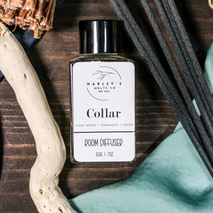 Op de afbeelding: Een kamergeurfles met een zwarte dop en een wit etiket met de tekst "Collar" en "Room Diffuser". De fles bevat 150g en is omgeven door zwarte diffuserstokjes, drijfhout en stof.
