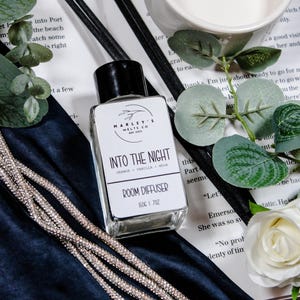 May include: A clear glass room diffuser with a black cap, labeled "Into The Night." The label lists orange, vanilla, and musk scents. Black reeds and decorative elements surround the bottle, including a white ceramic bowl and a silver beaded strand.