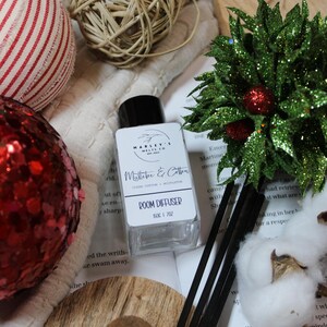Może przedstawiać: Przezroczysta szklana butelka dyfuzora do pomieszczeń z czarną nakrętką, z napisem "Mistletoe & Cotton". Obraz zawiera elementy dekoracyjne, takie jak czerwona błyszcząca ozdoba, zielona błyszcząca roślina i drewniane kule. Dyfuzor ma pojemność 150 ml.