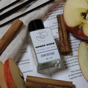 May include: A clear glass room diffuser bottle with a black cap, labeled "Spiced Cider." The image includes cinnamon sticks, apple slices, and black diffuser sticks, creating a cozy, autumnal theme. The bottle contains 150g or 7oz of product.