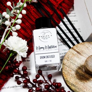 Może przedstawiać: Przezroczysta szklana butelka dyfuzora do pomieszczeń z czarną nakrętką, oznaczona jako "Merry Mistletoe". Etykieta zawiera nazwę marki "Marley's Melts Co." i opis zapachu. Obraz zawiera czerwone jagody, białe kwiaty i czarne patyczki dyfuzora.