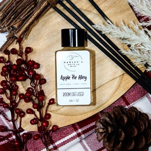 Może przedstawiać: Butelka dyfuzora do pomieszczeń z czarną nakrętką i białą etykietą z napisem "Apple Pie Alley". Butelka stoi na drewnianej tacy z elementami dekoracyjnymi, w tym jagodami, trzciną i szyszką. Butelka zawiera 28g płynu.