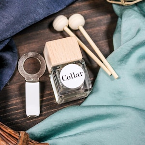 Könnte beinhalten: Eine klare Glasflasche mit einem Holzdeckel und einem weißen Etikett mit der Aufschrift "Collar". Die Flasche befindet sich neben einem silbernen Flaschenöffner und zwei Holzstäbchen mit weißen Kugelenden. Der Hintergrund umfasst blauen und türkisfarbenen Stoff.