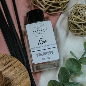 Puede incluir: Un difusor de ambiente de vidrio transparente con tapa negra y etiqueta blanca que dice "Eve" y "Difusor de ambiente". La botella contiene 150g y está rodeada de palitos difusores negros, esferas decorativas y vegetación.