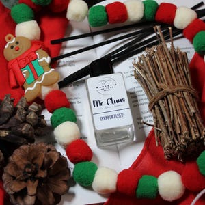 Może przedstawiać: Świąteczna aranżacja z dyfuzorem do pomieszczeń "Mr. Claus", ozdobą w kształcie piernikowego ludzika oraz girlandą z czerwonych, zielonych i białych pomponów. W zestawie również szyszki, patyczki do dyfuzora i pęczek gałązek.