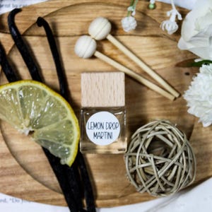 Könnte beinhalten: Auf einem Holztablett befindet sich eine kleine Glasflasche mit Holzverschluss, beschriftet mit "LEMON DROP MARTINI". Dazu Vanilleschoten, eine Zitronenscheibe, dekorative Stäbe und ein geflochtener Ball. Weiße Blumen und Text im Hintergrund.
